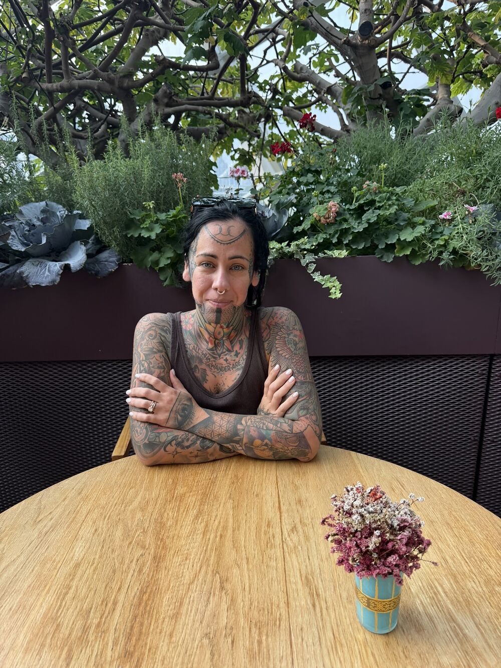 A tattooed woman seated at a wooden garden table with a small vase of flowers