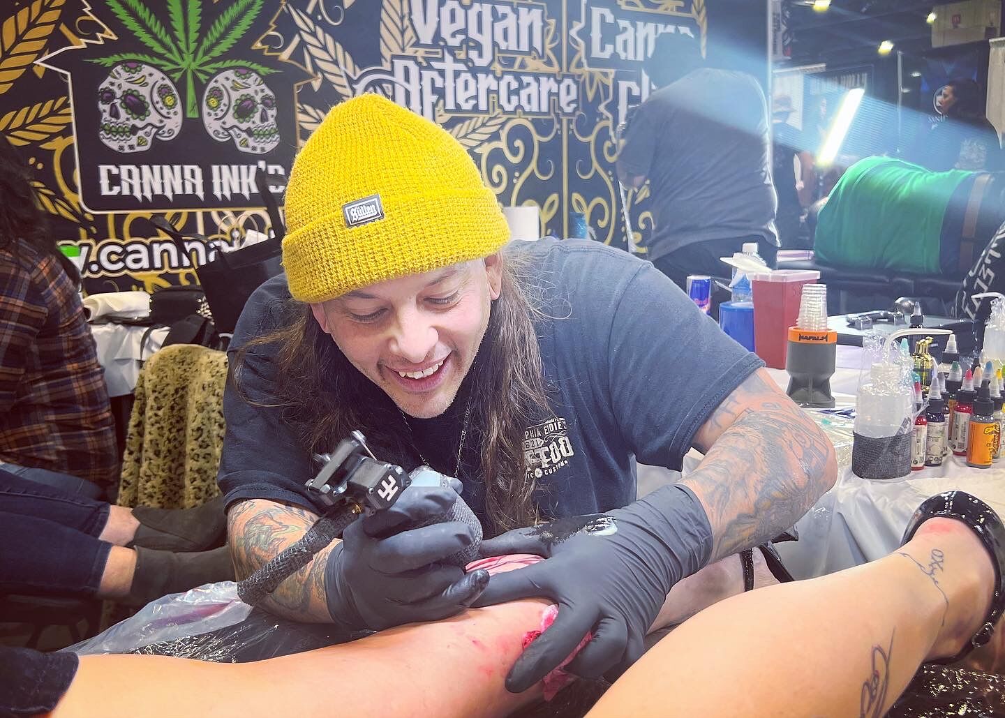 A tattoo artist in a yellow beanie working on a client's arm at a convention booth